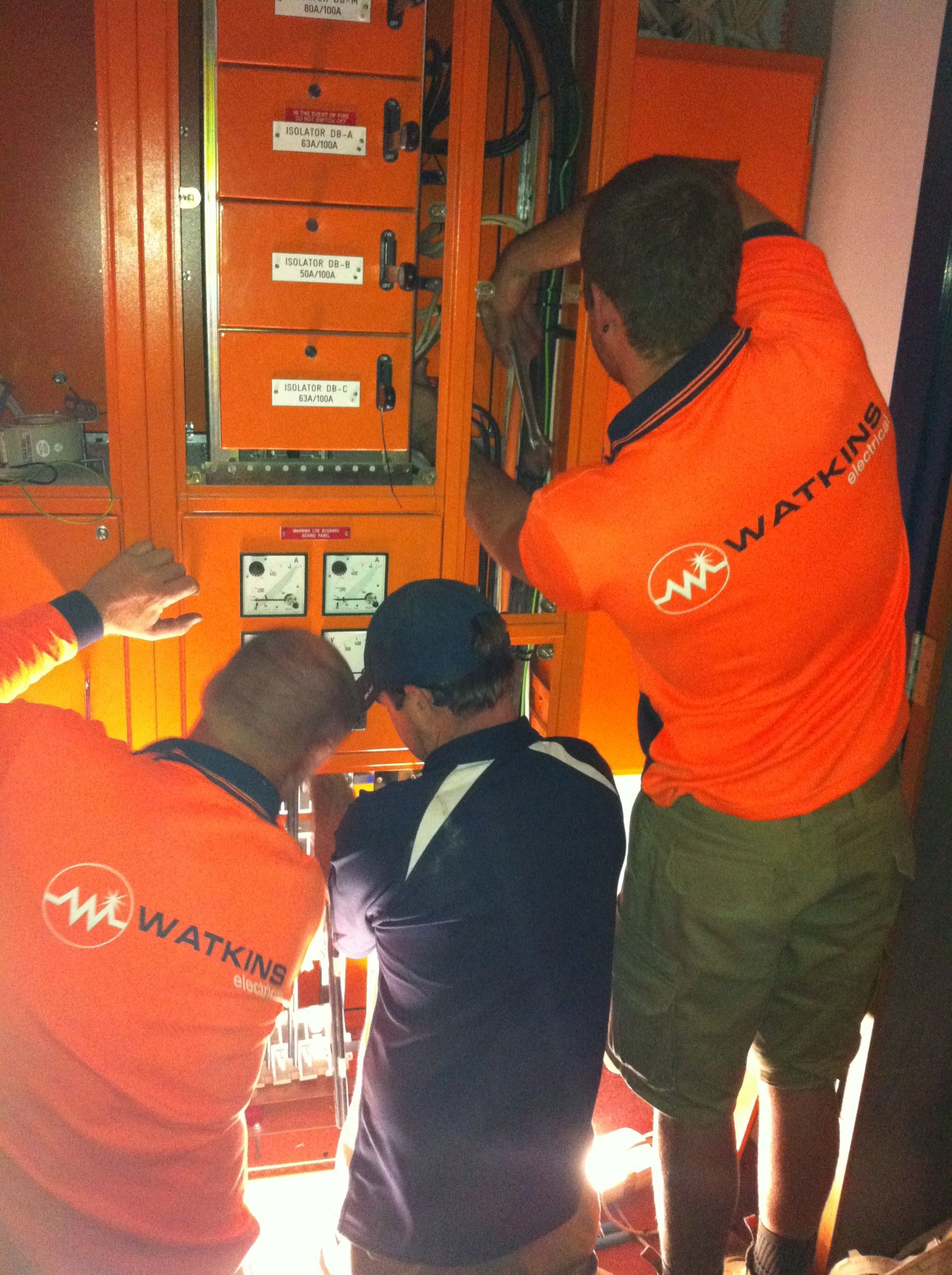 Three electricians in orange shirts working on an orange electrical panel — Watkins Electrical In Pialba, QLD
