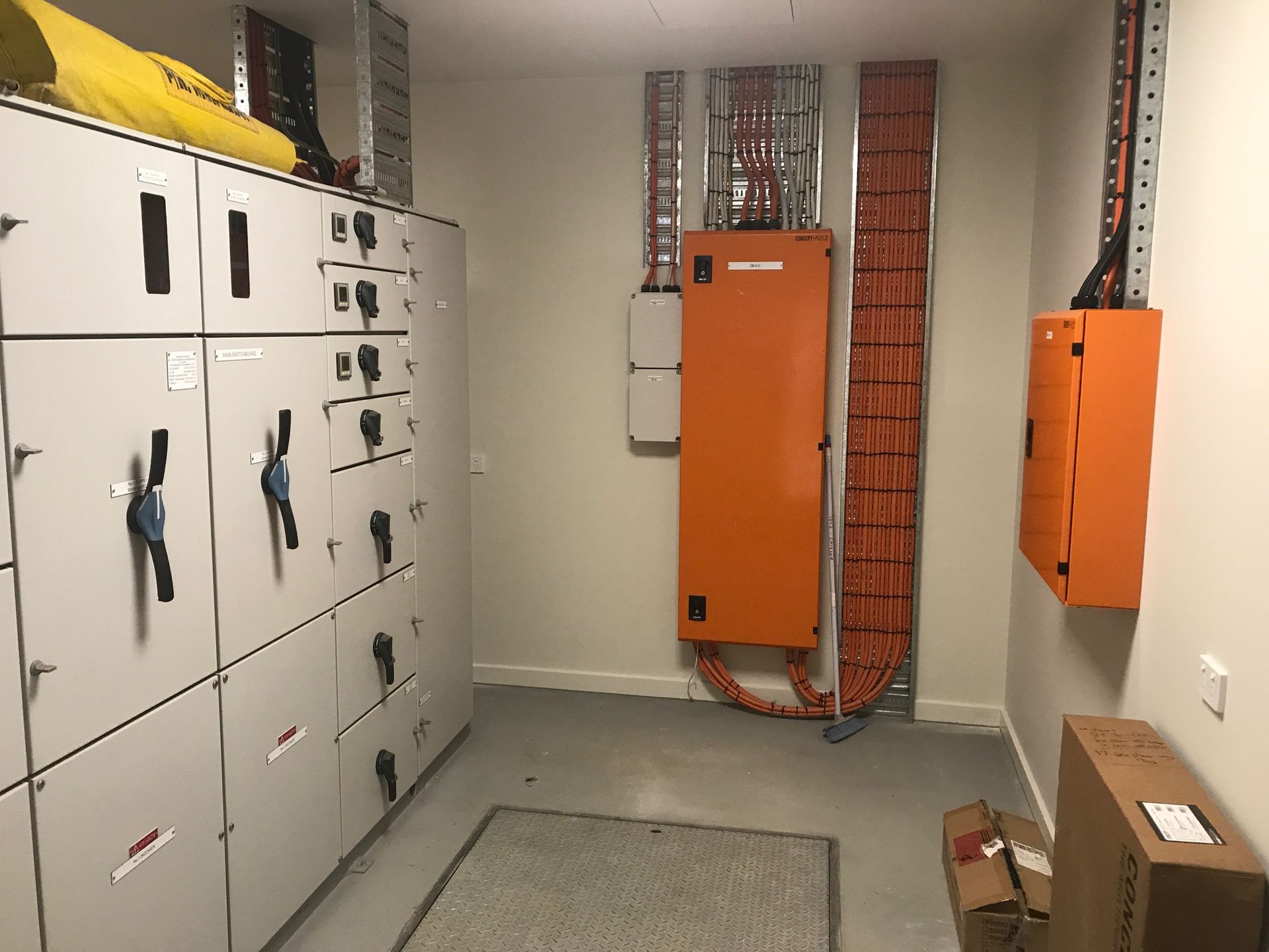 Electrical room with white cabinets, orange panels, and conduit against beige walls, and a gray floor — Watkins Electrical In Pialba, QLD