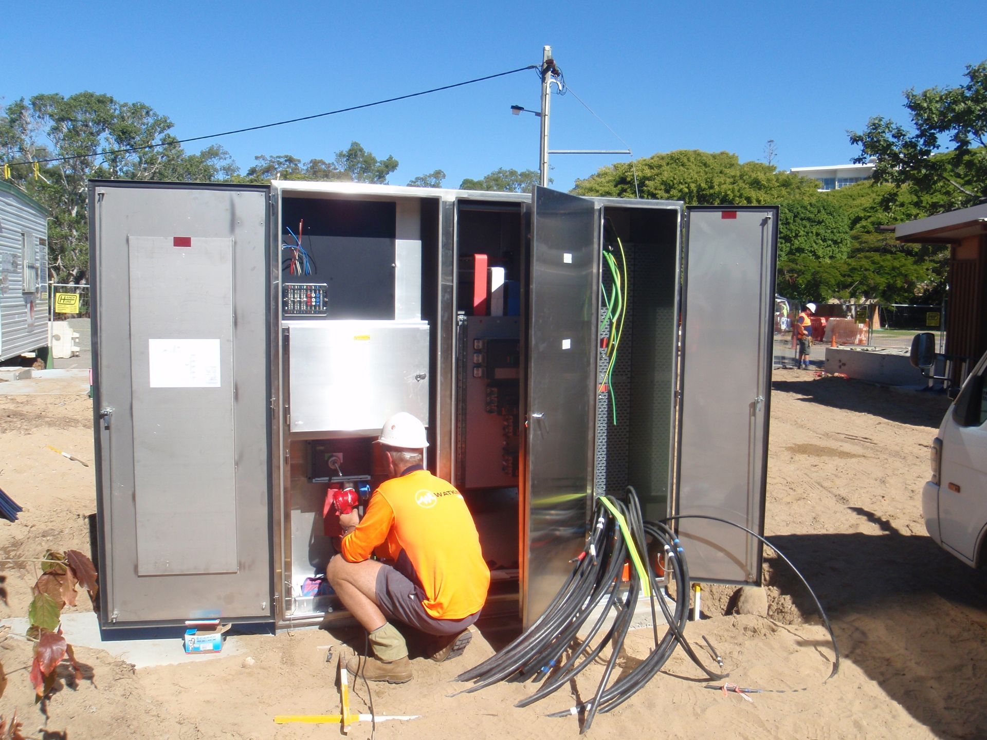 A worker in an orange shirt and hard hat works on electrical equipment in an outdoor cabinet — Watkins Electrical In Pialba, QLD