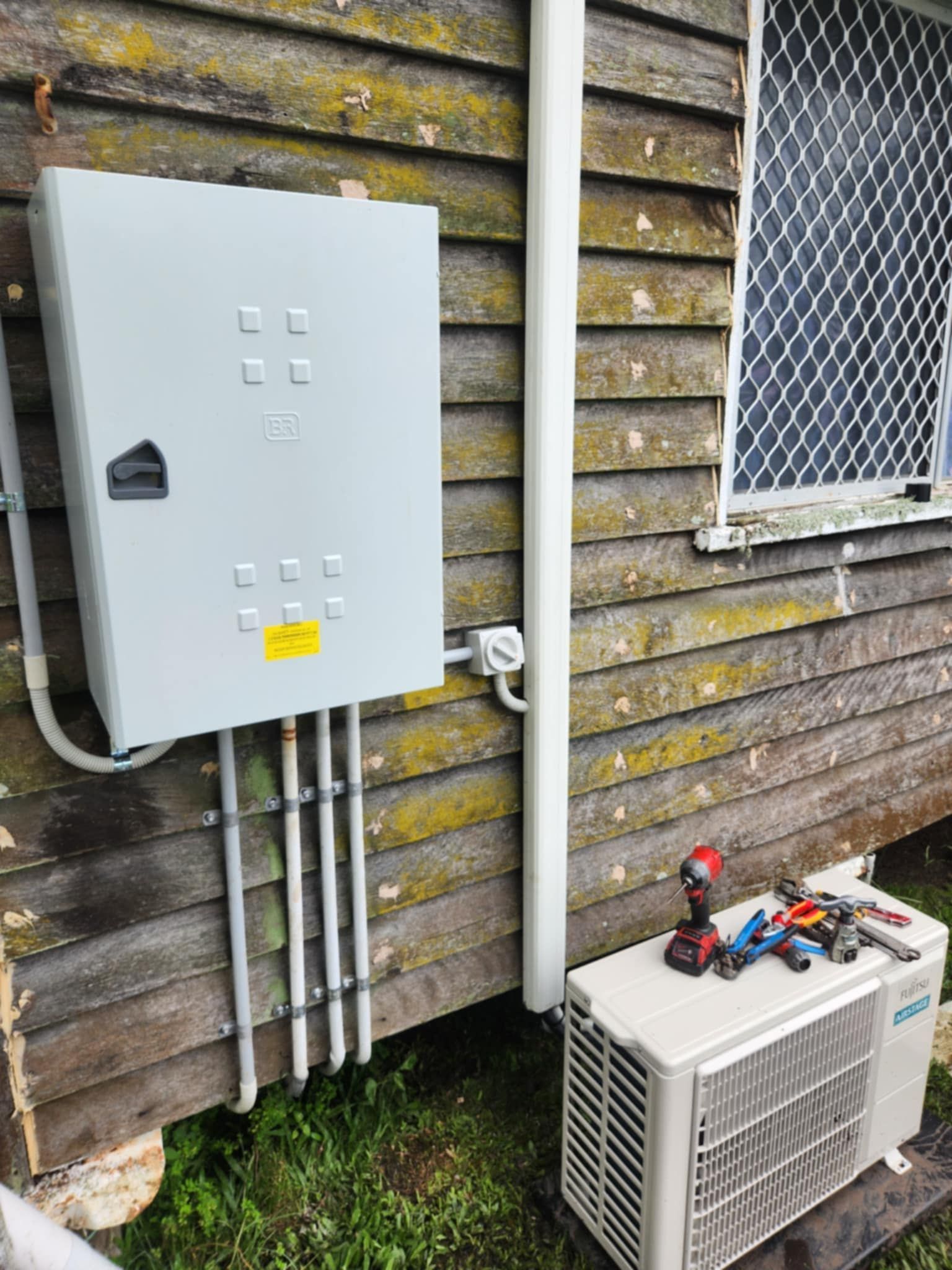 Electrical panel and air conditioner installed on weathered wooden building — Watkins Electrical In Pialba, QLD