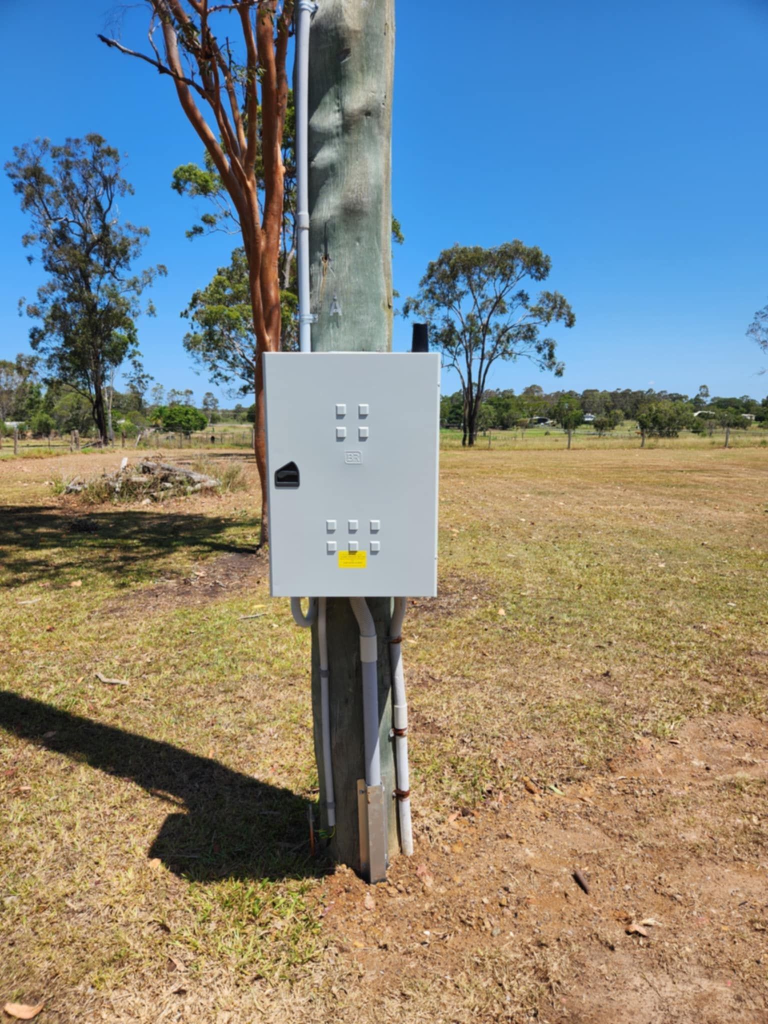 Grey electrical box mounted on a wooden utility pole in a grassy field under a clear blue sky — Watkins Electrical In Maryborough, QLD