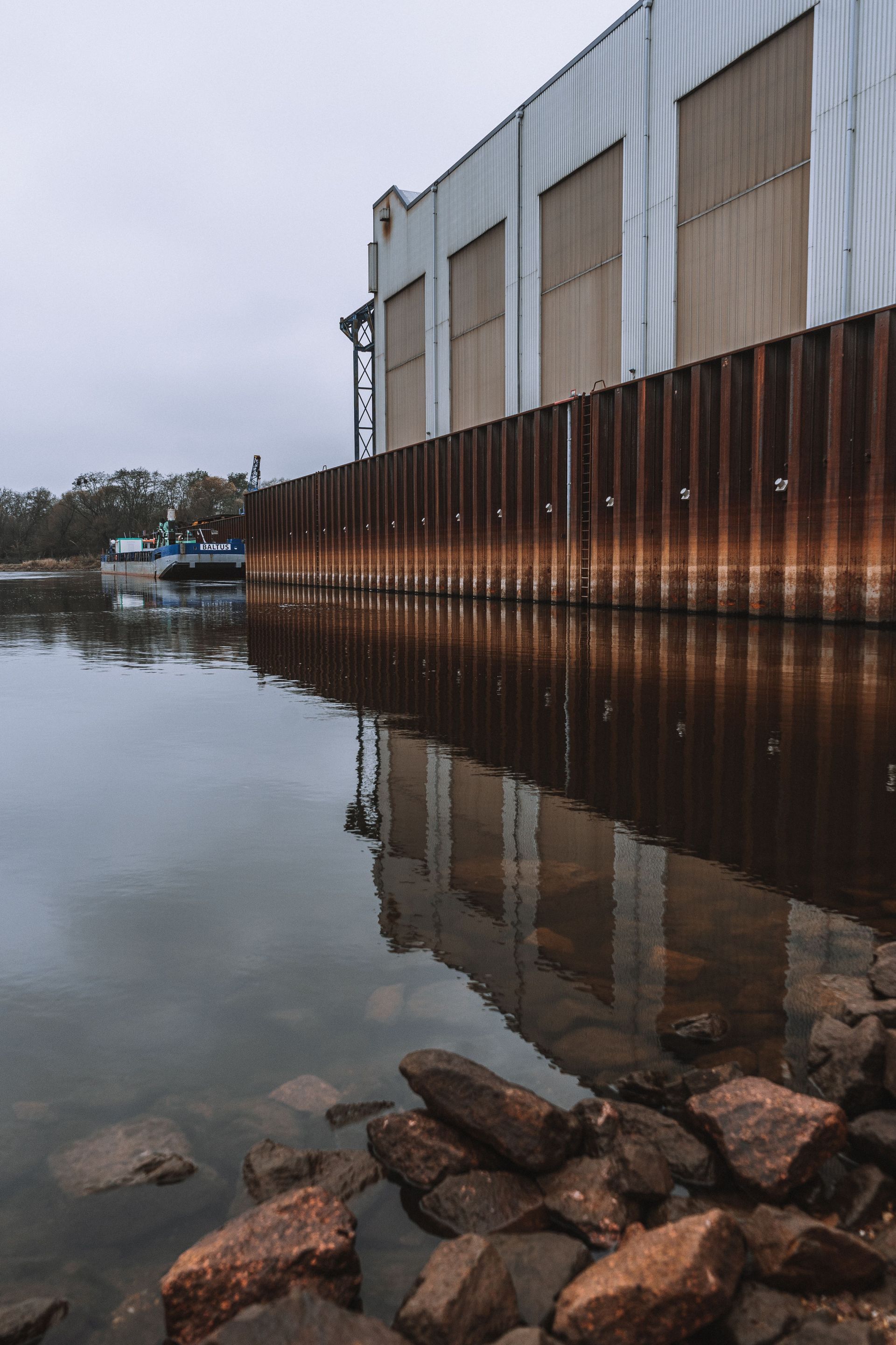 Rusty industrial wall beside calm water, with rocks in the foreground and reflections at dusk.