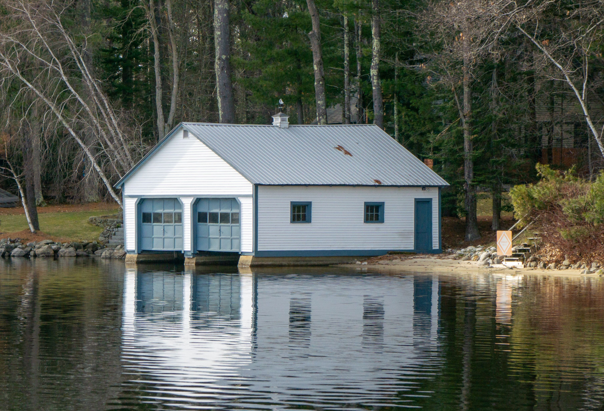 White boathouse by a wooded lake, with its reflection on calm water.