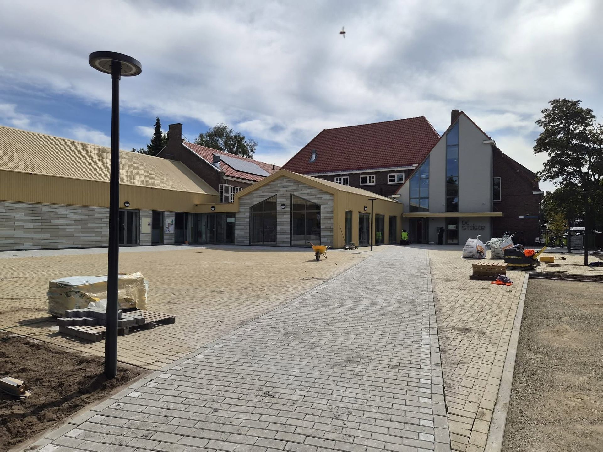 Moderne binnenplaats met een geplaveid pad, lantaarnpalen, banken en lage gebouwen onder een bewolkte hemel.
