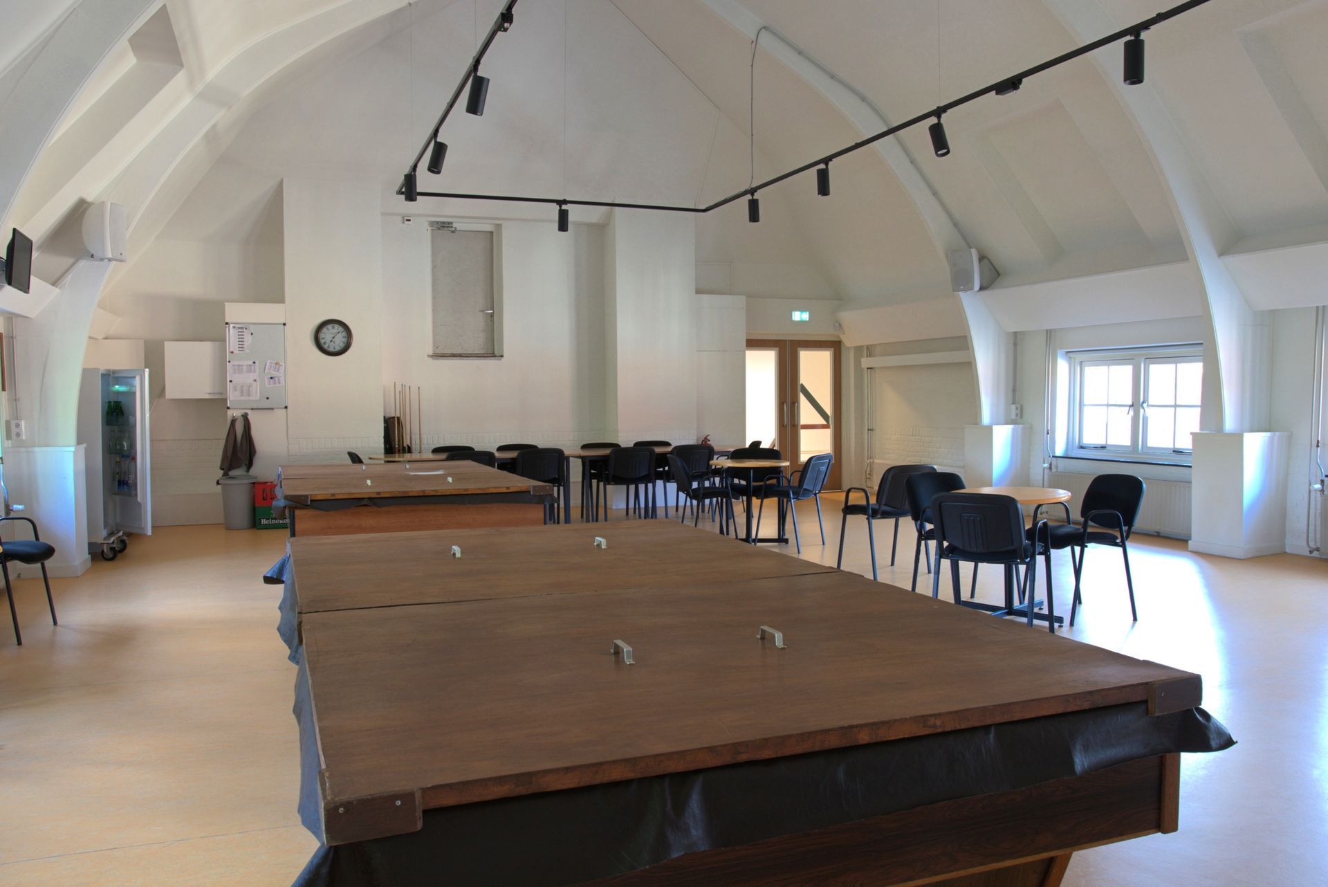 Een lege gemeenschapszaal met tafels en stoelen onder een hoog, wit gewelfd plafond.