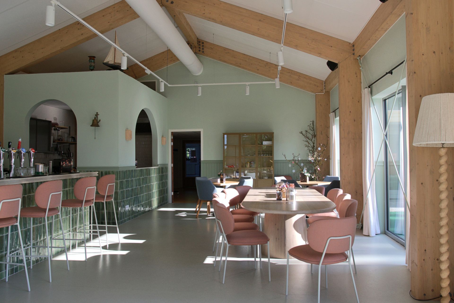 Lichte café-eetzaal met roze stoelen, tafels en hoge houten balken, verlicht door grote ramen.