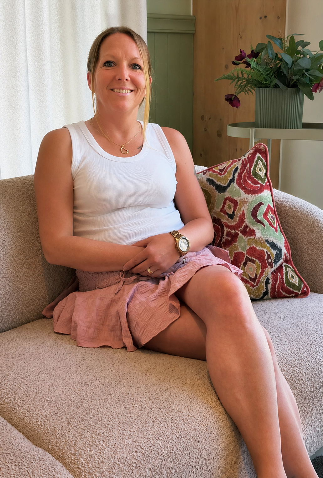 Vrouw zit op een bank in een lichte woonkamer, gekleed in een witte top en een roze rok, en lacht naar de camera.