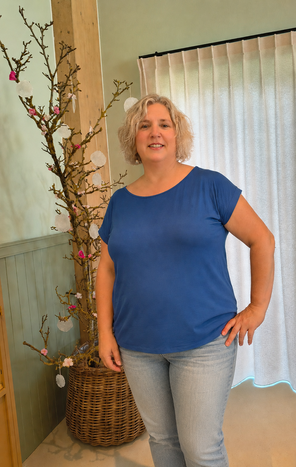 Vrouw in een blauwe blouse en spijkerbroek staat binnen naast een decoratief arrangement van takken en een gordijn.