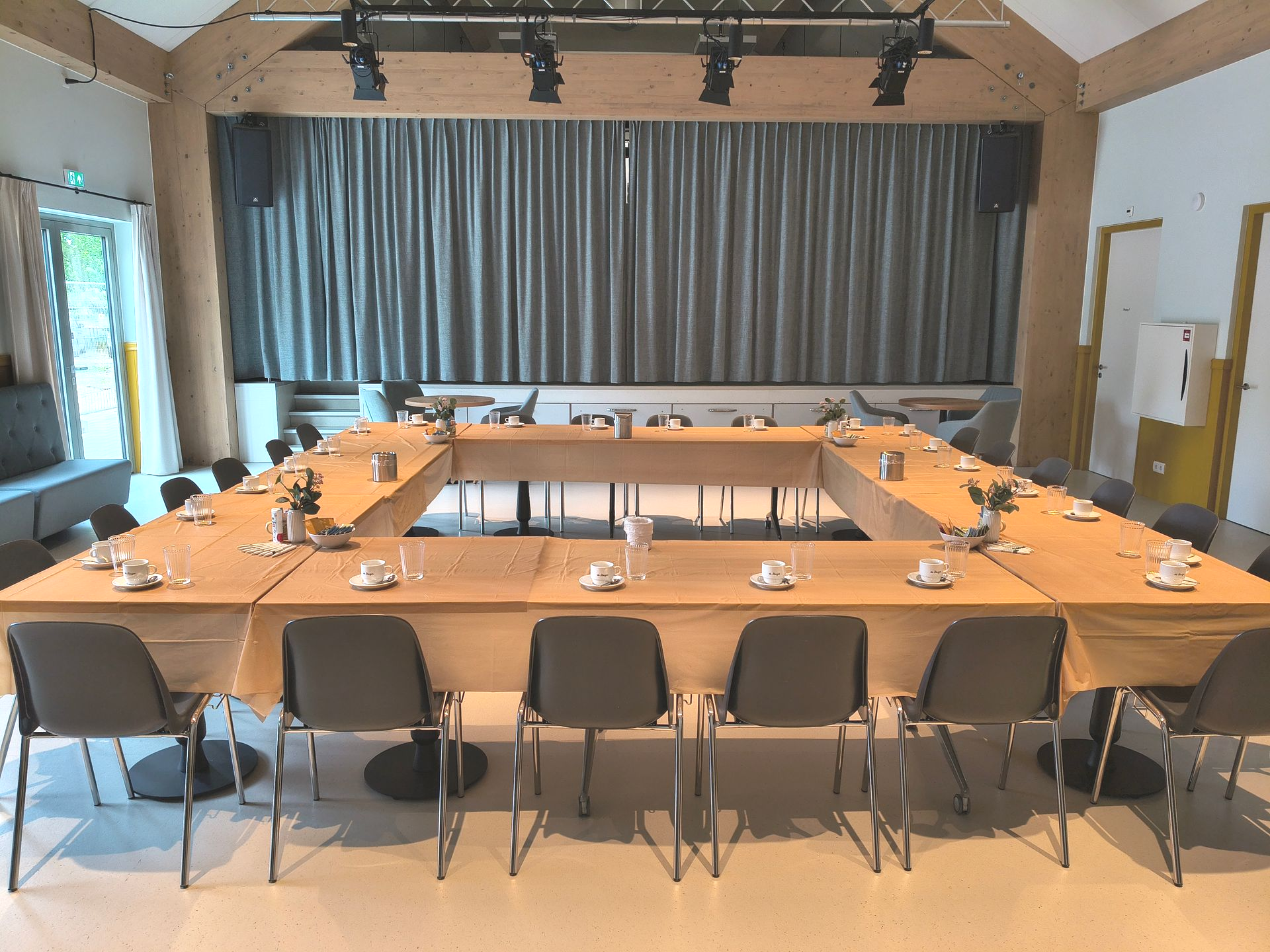 Vergaderruimte met een  tafel, stoelen, microfoons en een gesloten gordijn aan de achterwand.