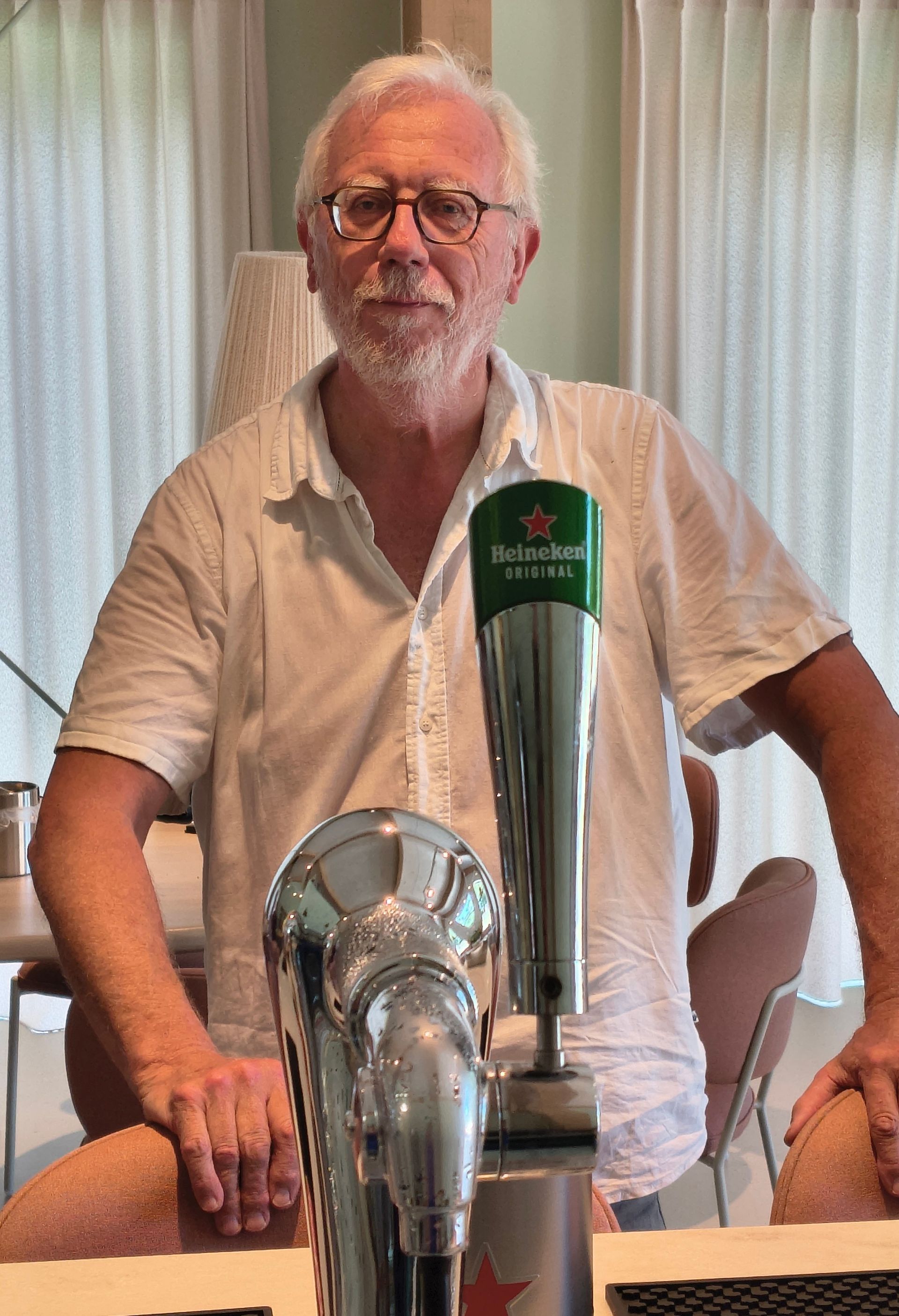 Een man staat achter een biertap met een groene Stella Artois-hendel, binnen bij een raam en gordijnen.