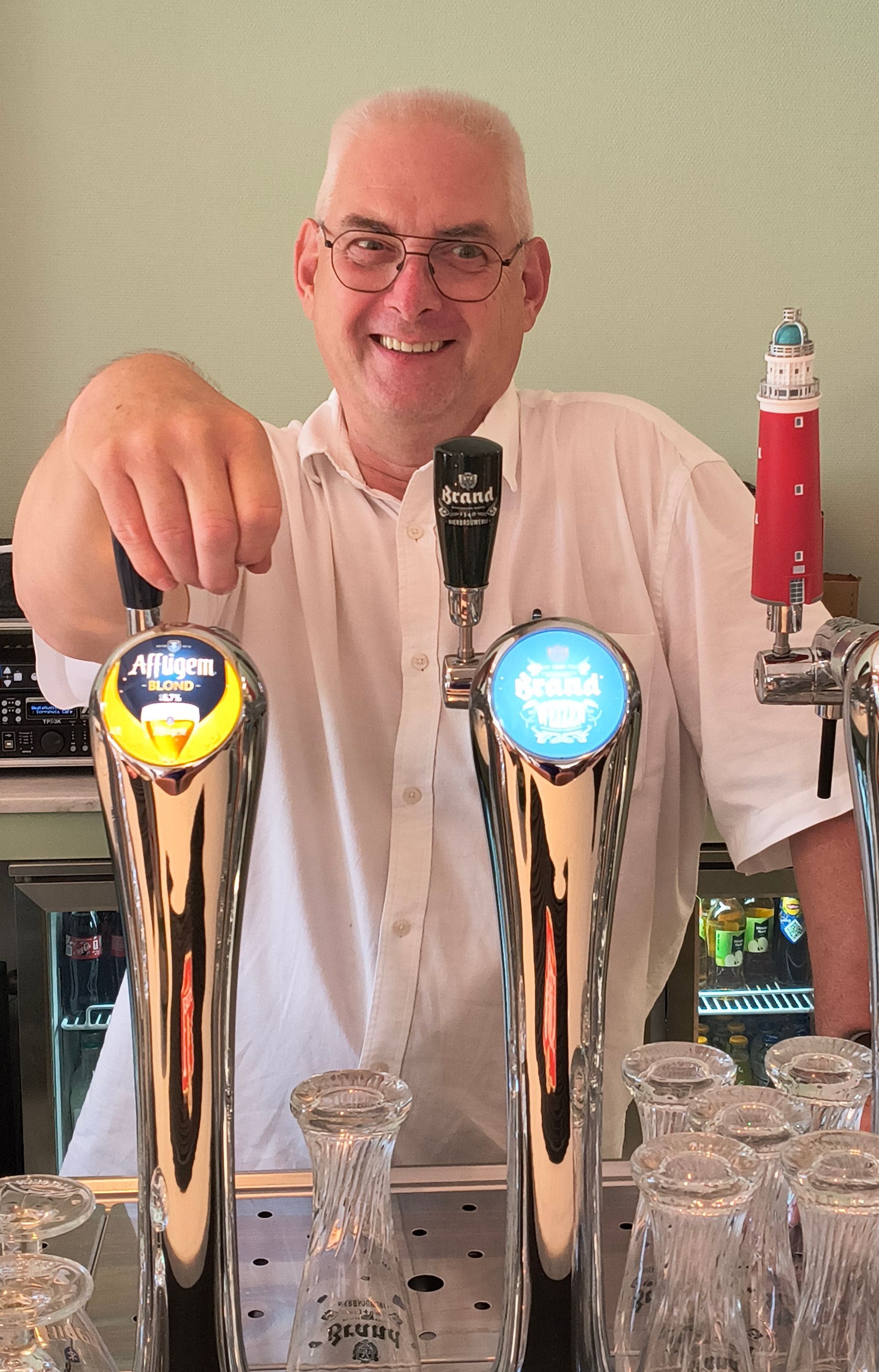 Een barman staat lachend achter twee biertaps met rode en blauwe taphendels aan de bar.