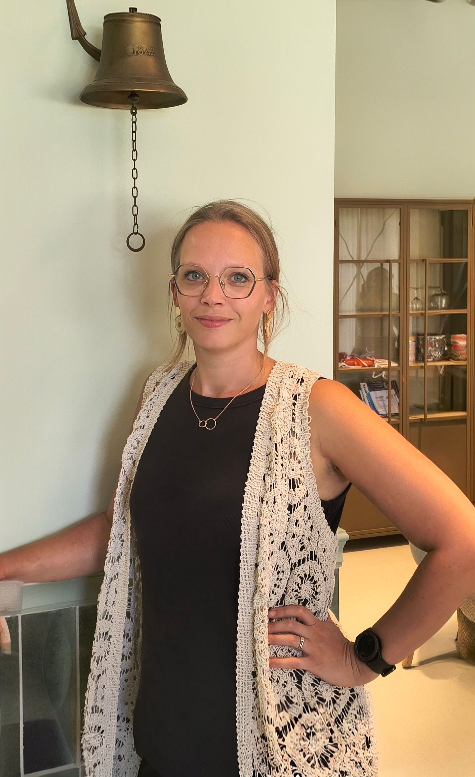 Vrouw staat binnen naast een bel, gekleed in een zwarte top en een wit vest, hand in haar zij, lachend naar de camera.