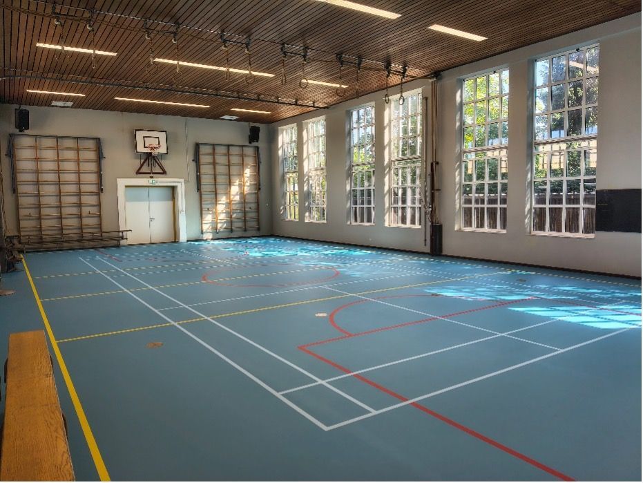 Een lege gemeenschapszaal met tafels en stoelen onder een hoog, wit gewelfd plafond.