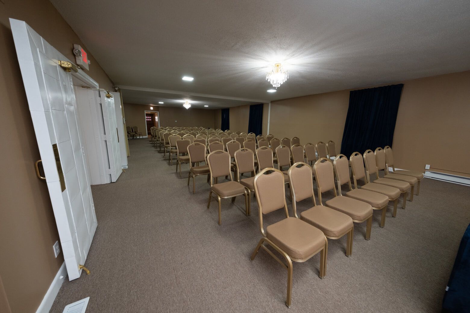 Empty conference room with rows of tan chairs, white walls, carpeted floor, and overhead lights.