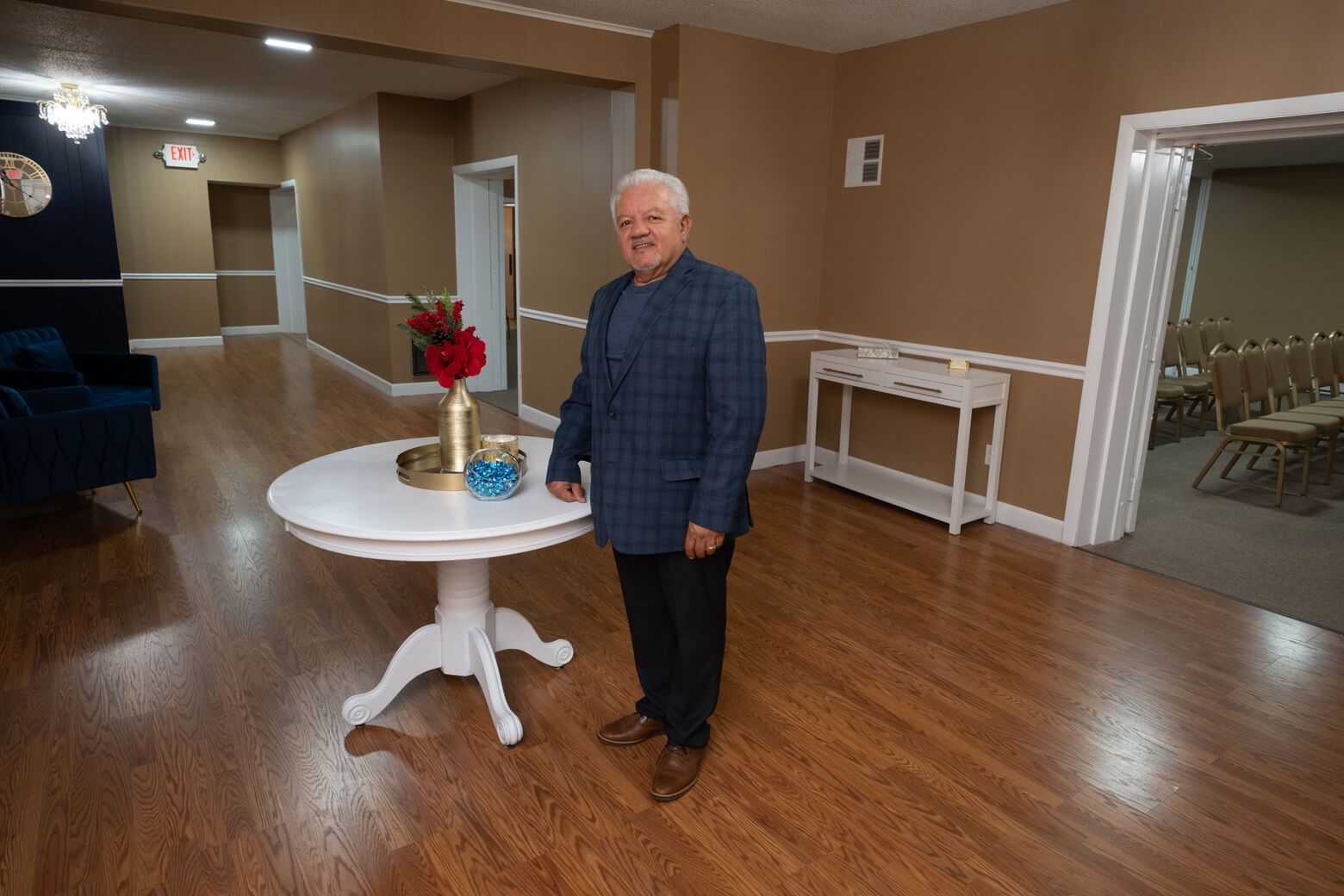 Man standing beside a white table with a red flower arrangement in a bright hall with hardwood floors