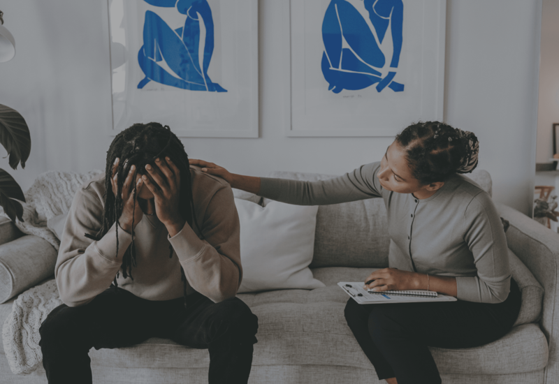 Two people on a couch; one sits with head in hands while the other comforts them.