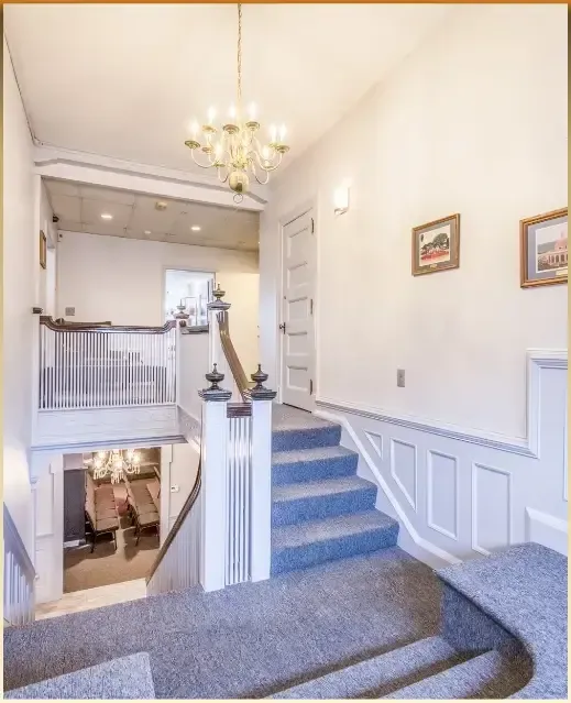 Bright stairwell with blue carpet, white wainscoting, chandelier, and framed wall art in a house foyer