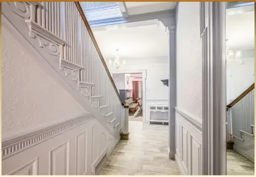 Bright hallway with white wainscoting, staircase, and a view into a light-filled room with a sofa.