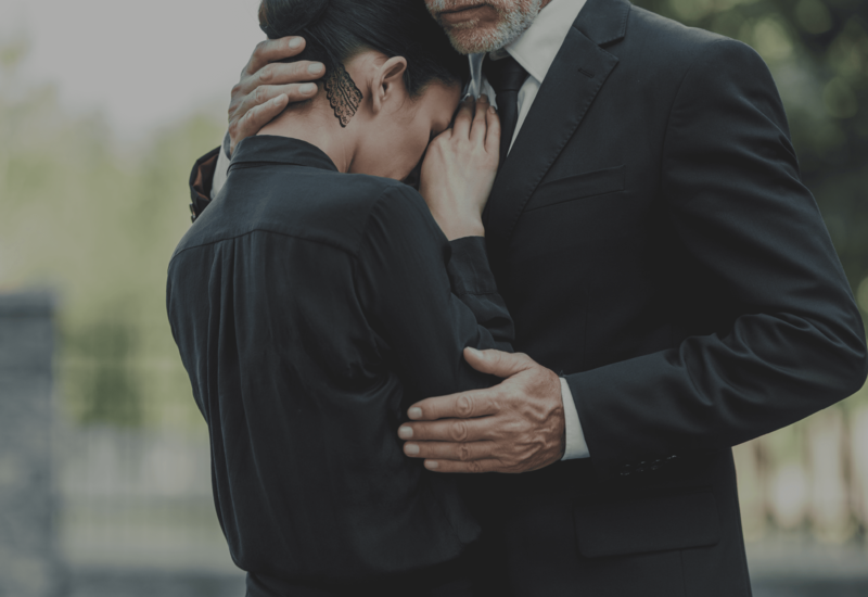 Two people in black clothing embrace closely outdoors, one comforting the other.