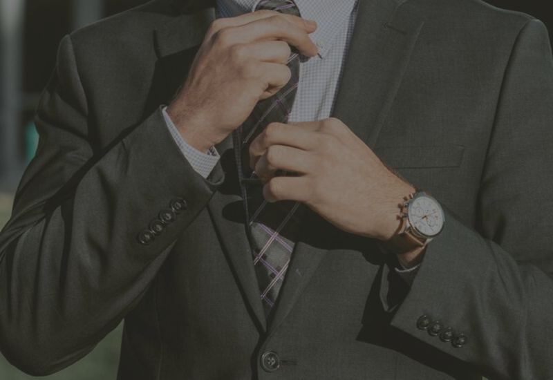 Man in a dark suit adjusting his tie and buttoning his jacket, wearing a wristwatch.