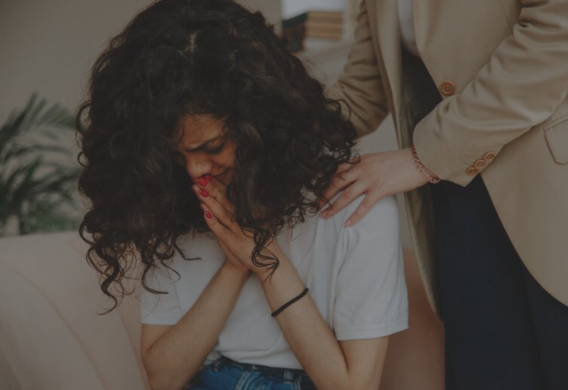 Woman sitting with head in hands while another person comforts her with a hand on her shoulder