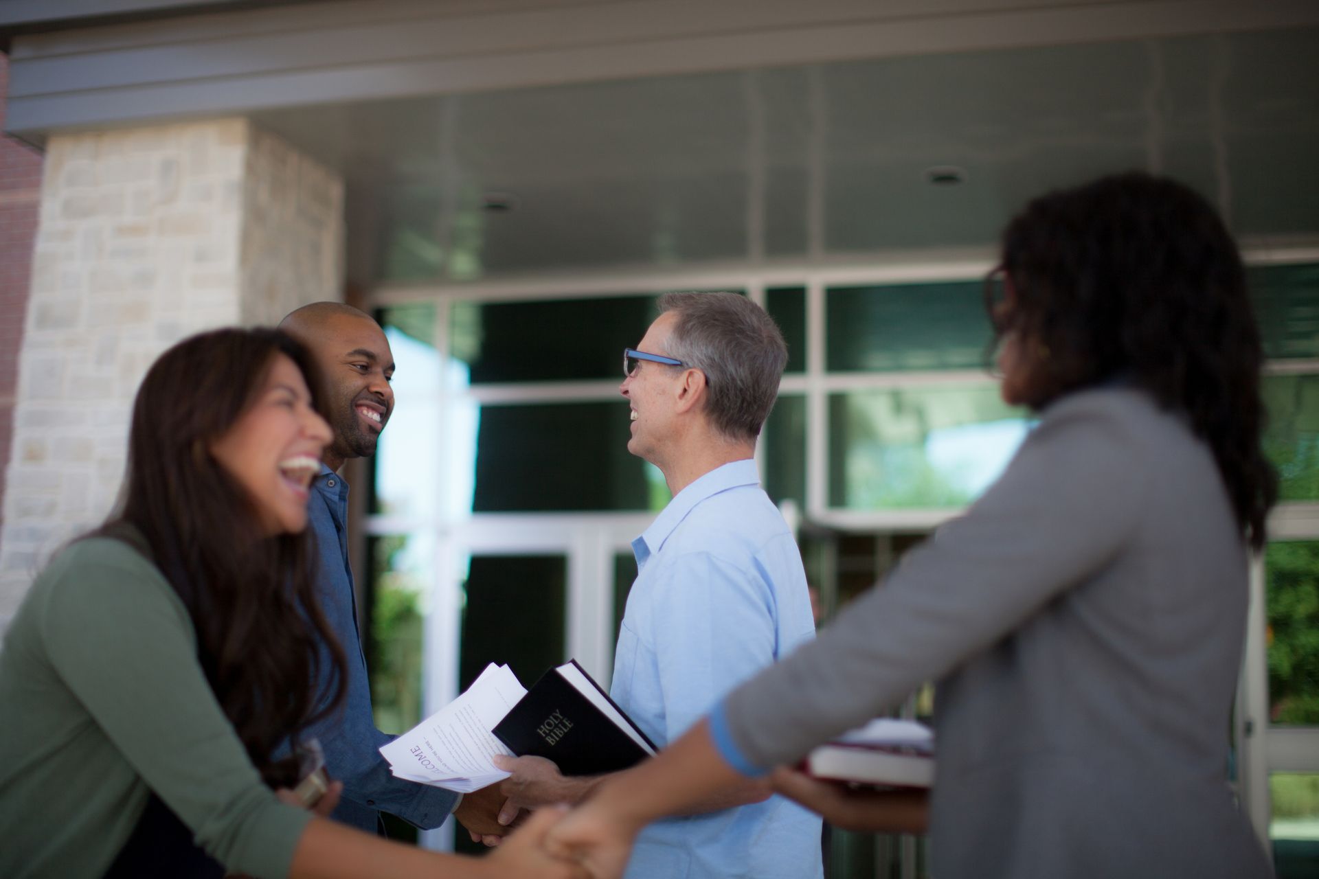 People greeting and shaking hands outside a building, smiling and holding folders.