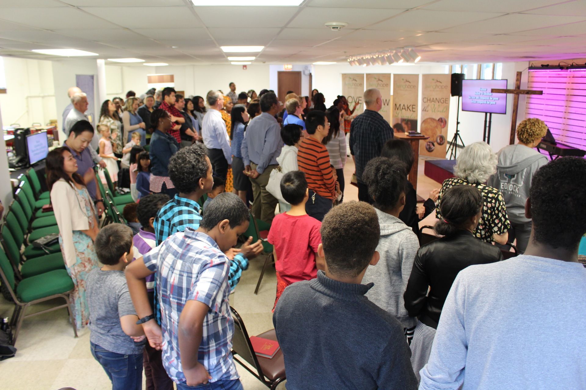 Crowded hallway gathering with people standing and talking near tables and a pink-lit screen.