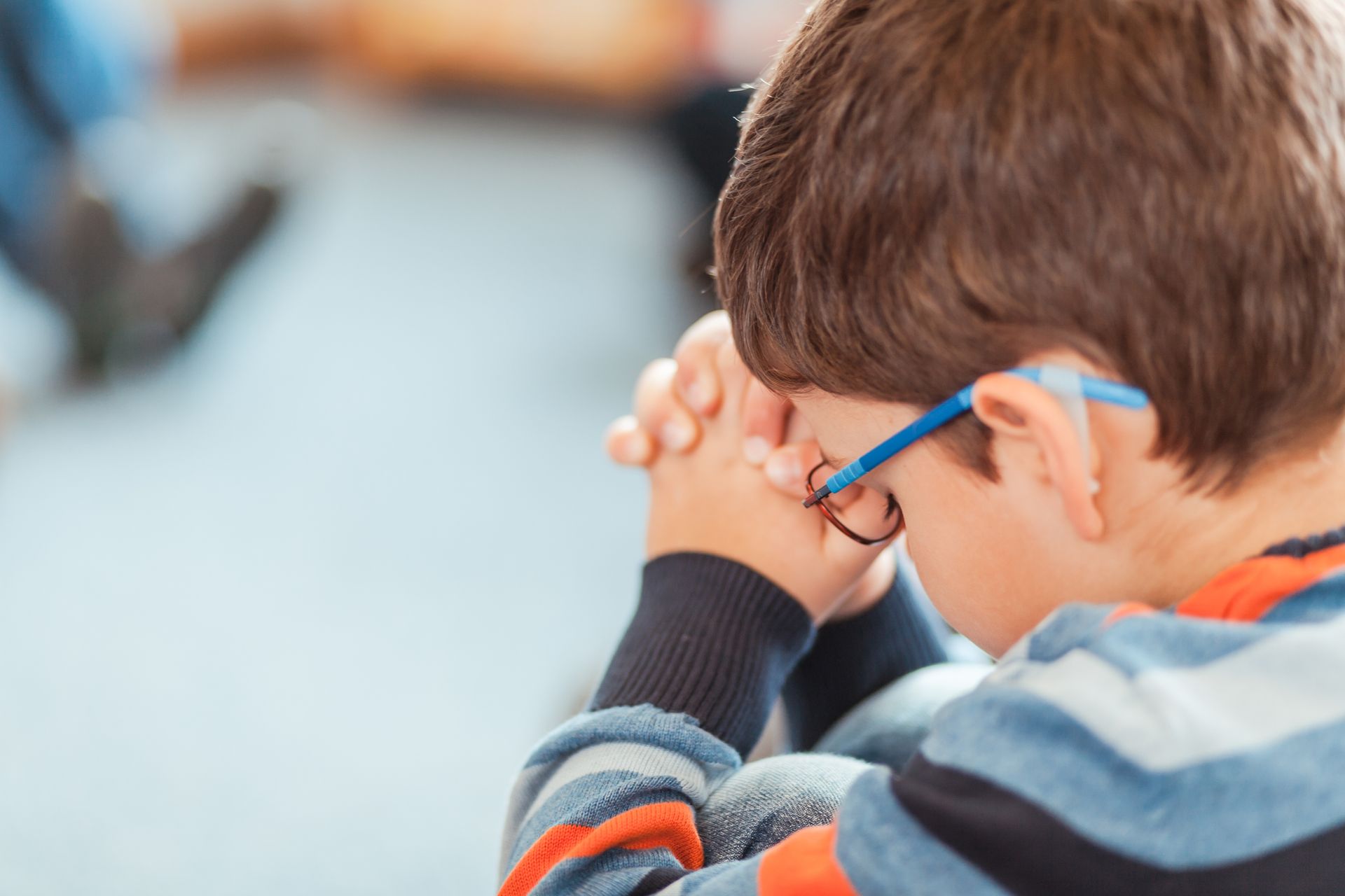Child with blue glasses resting clasped hands near face in a blurred indoor setting