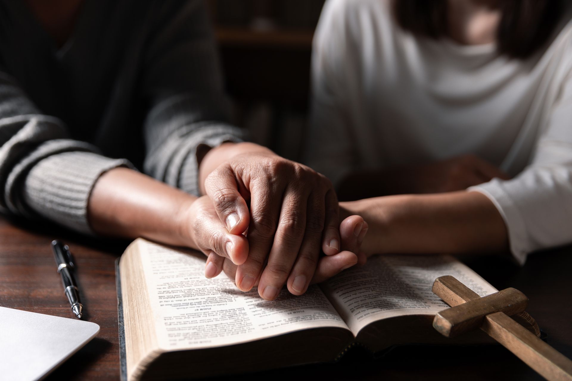Two people hold hands over an open book beside a wooden cross on a table.