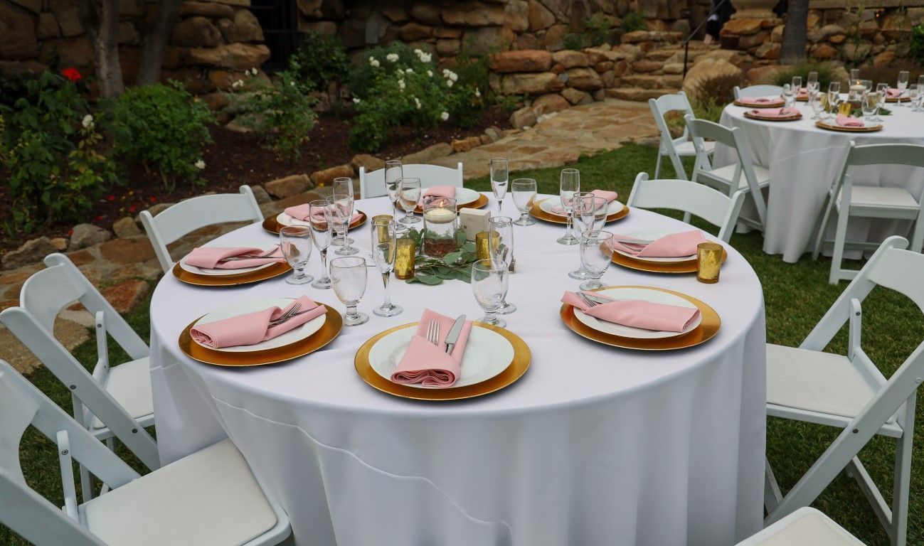 Round white-clothed banquet table with pink place settings and floral centerpiece in a garden setting