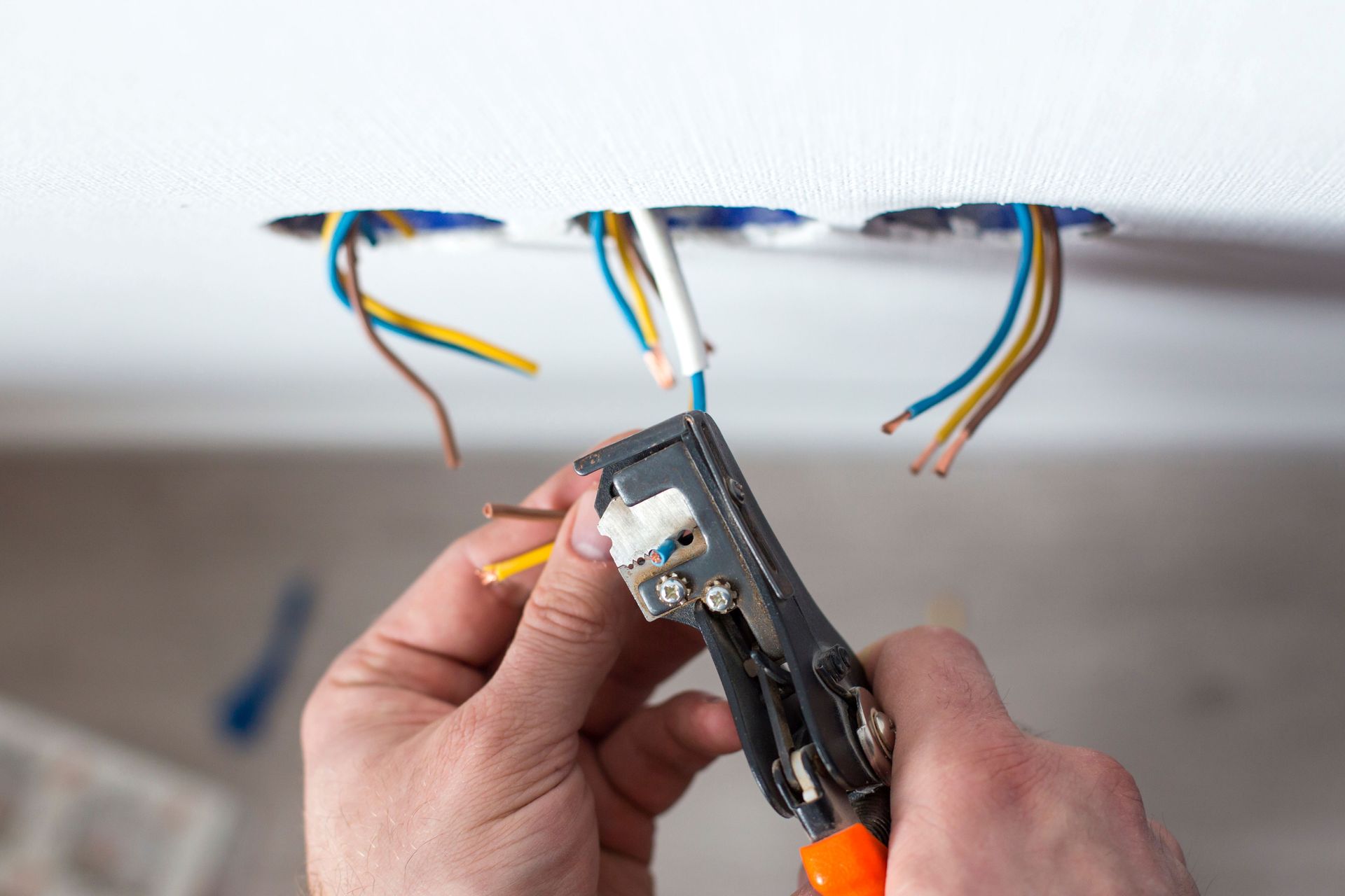 Hands strip insulation from exposed electrical wires in a ceiling opening using a wire stripper.