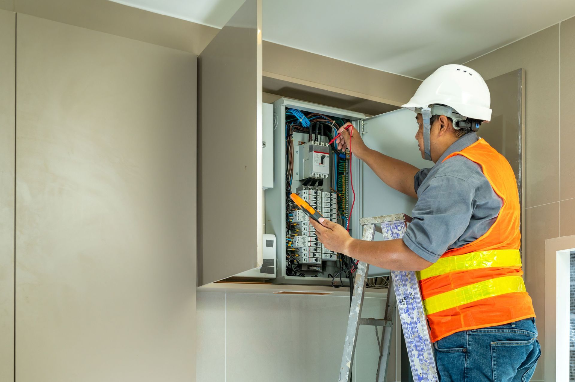 Electrician in hard hat and safety vest working inside an open electrical panel on a ladder.
