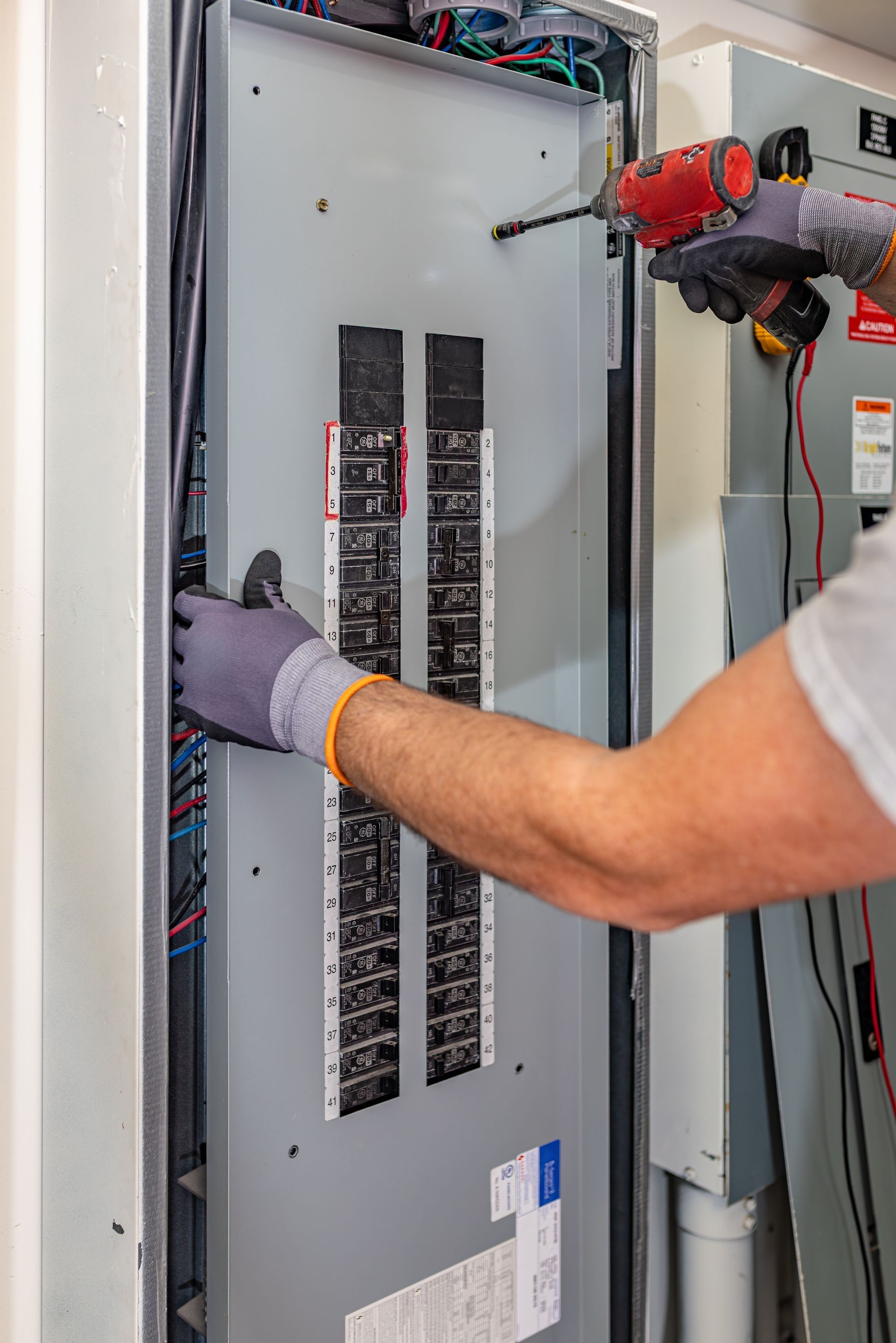 Technician in hard hat inspecting electrical panel with a multimeter, wearing gloves and safety glasses.