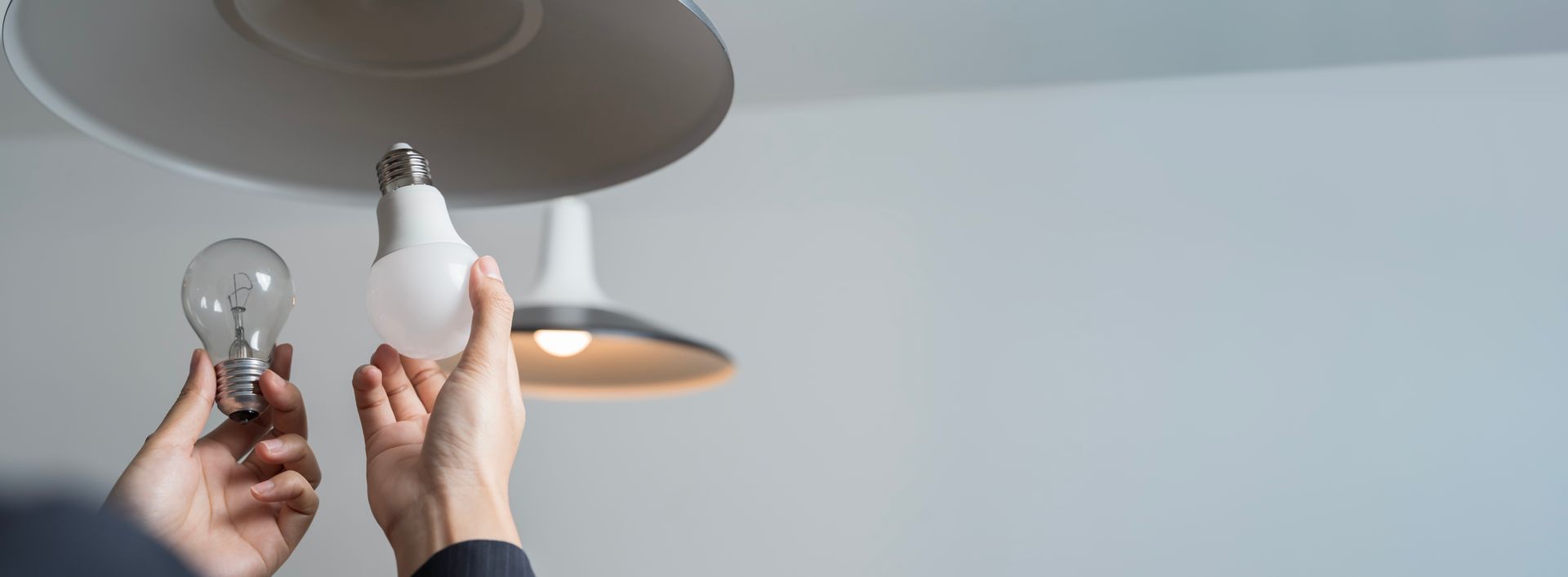 Hands holding a light bulb and a white LED bulb beneath a ceiling light fixture
