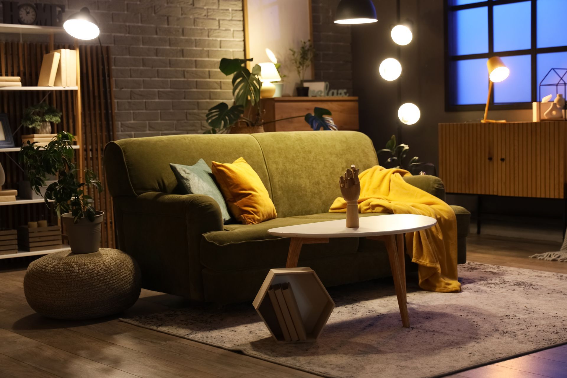 Cozy living room with green sofa, warm lighting, round table, plants, and shelves against a brick wall.