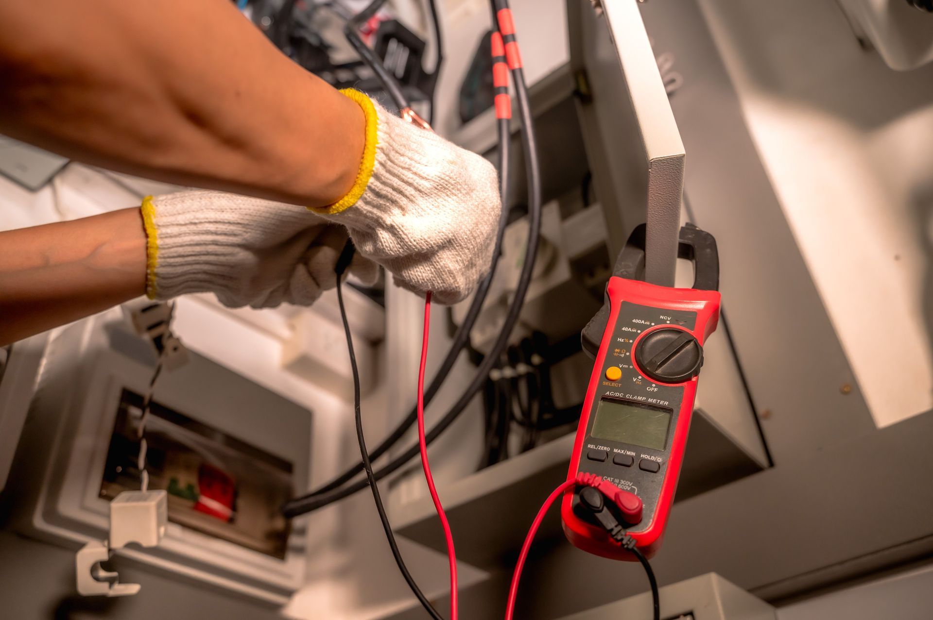 Gloved hands using a red multimeter to test wires inside an electrical panel