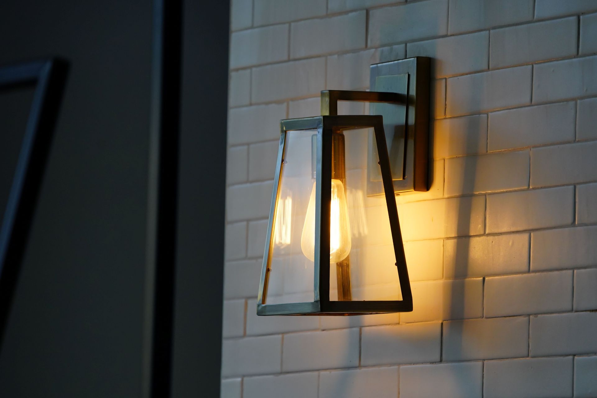 Wall-mounted lantern sconce glowing warm light against a white tiled wall