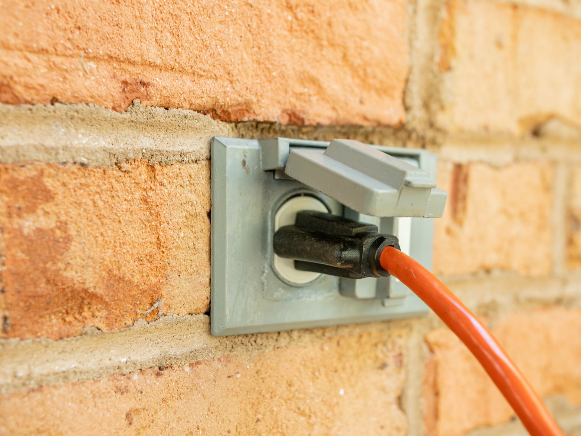 Orange extension cord plugged into an outdoor wall outlet on a brick wall.