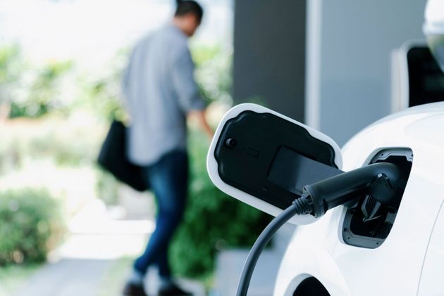Electric vehicle charging plug connected to a white car, with a person walking in the background.
