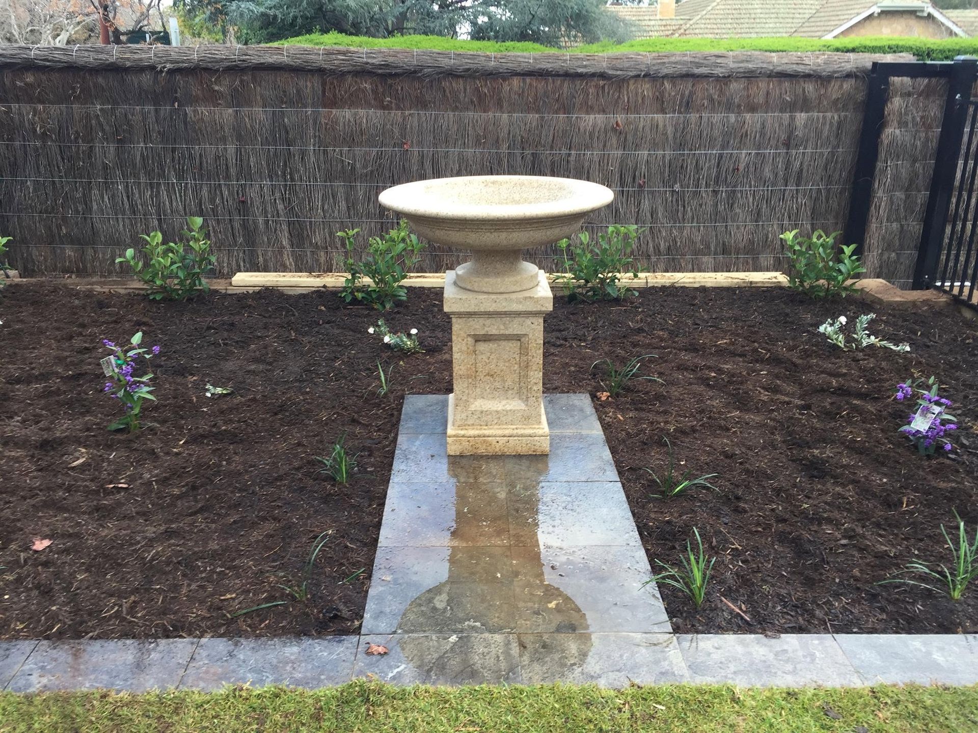 Garden birdbath centered on a stone path between freshly planted flower beds and a brick wall