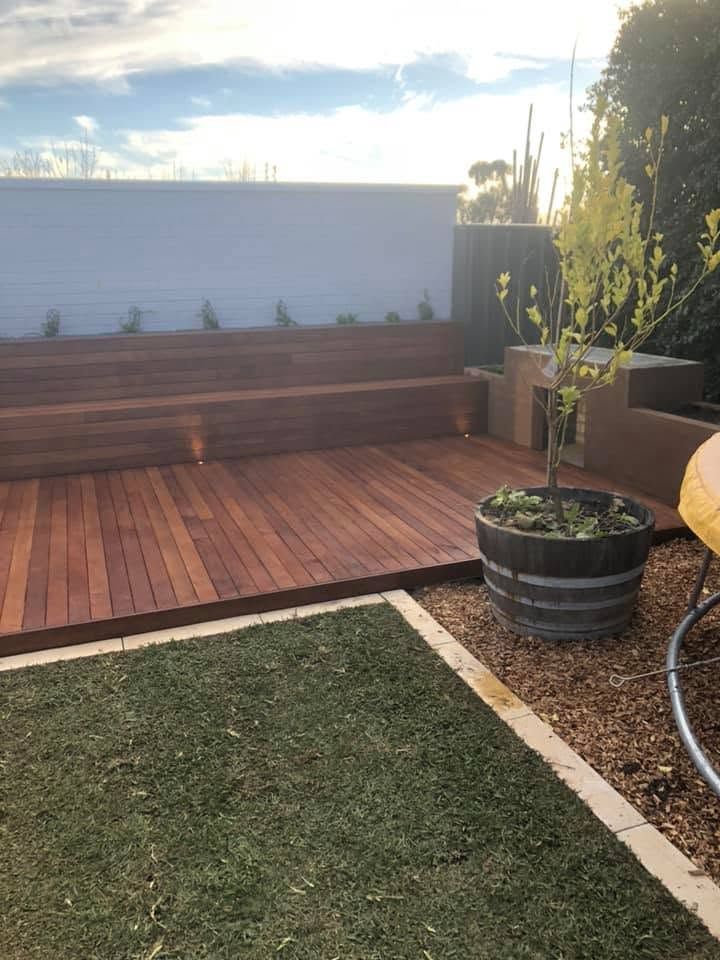 Backyard patio with wooden deck, bench seating, potted plants, and a grassy lawn in warm sunlight
