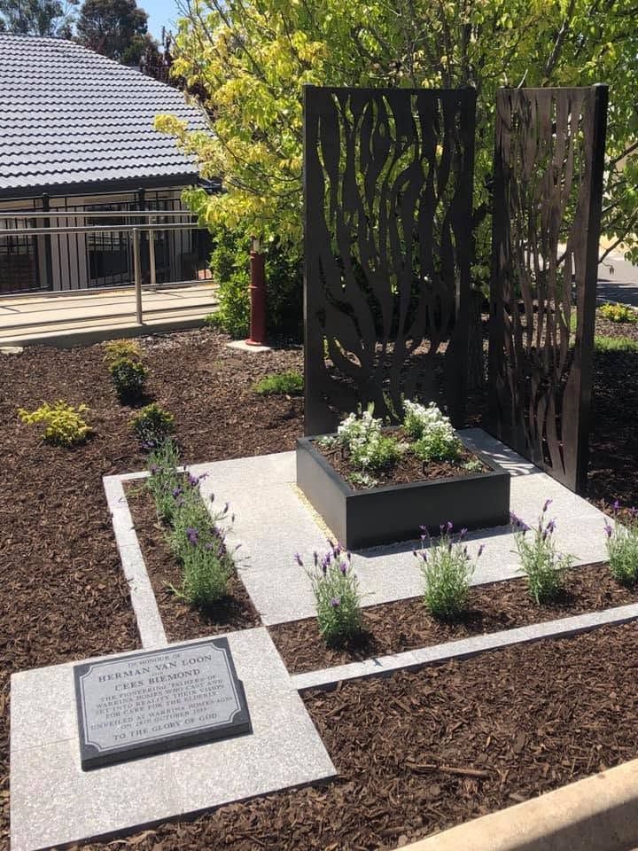 Outdoor memorial garden with a black metal sculpture, plaques, and planted beds beside a building.