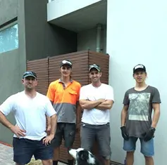 Four men standing outdoors in front of a building, with a dog at their feet.
