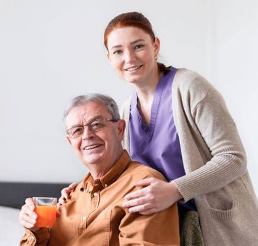 A woman in purple scrubs and cardigan smiles as she supports a man holding juice. Bright, indoor setting.