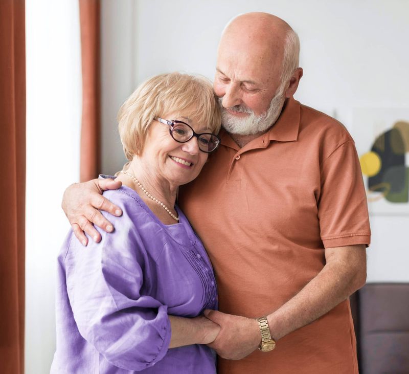 Smiling couple embracing indoors; man's arm around woman, holding hands.
