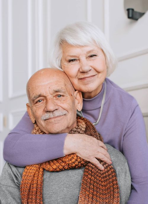 Elderly couple embracing, smiling. Woman's arms around man's neck. Man wearing scarf. White background.