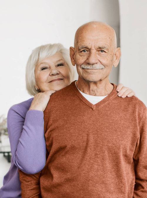 Elderly couple smiling, woman with hand on man's shoulder. Man wears rust-colored sweater.