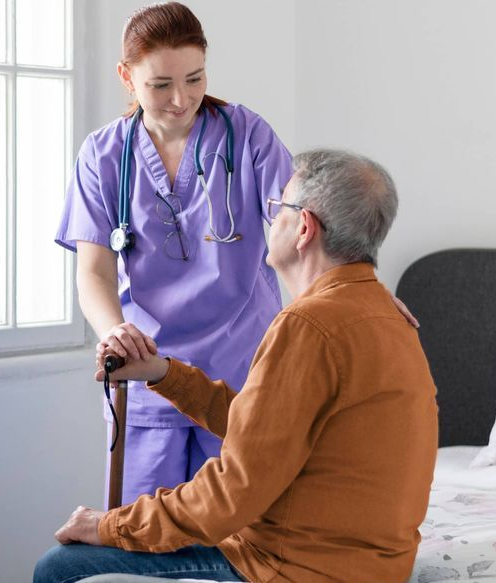 Nurse in purple scrubs assists a man, who is sitting, using a cane. They are in a room by a window.