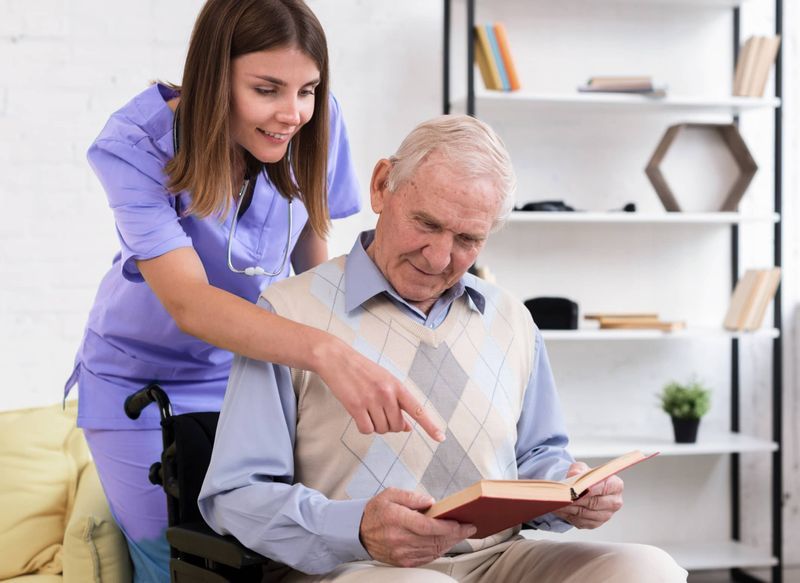 Nurse points to a book while assisting a senior man in a wheelchair; reading together indoors.