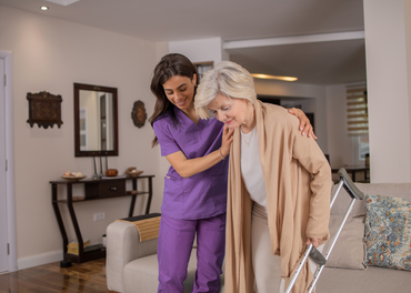 Caregiver assists an older person walking with a walker indoors.